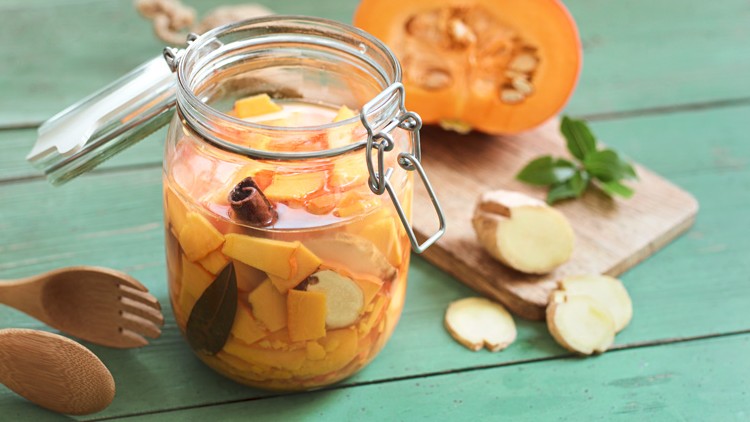 Ein Glas mit eingelegtem Kürbis, Ingwer und Gewürzen steht auf einem grünen Holztisch, daneben Kürbis und Ingwerscheiben.