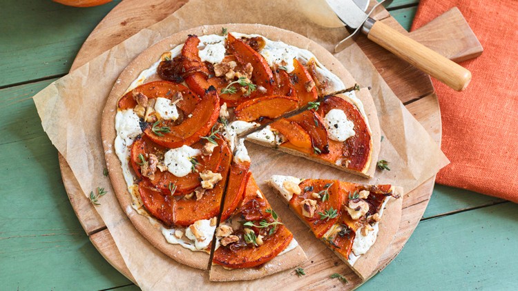 Runde Pizza mit Kürbisspalten, Ziegenkäse, Walnüssen und frischen Kräutern auf einem Holzbrett mit Backpapier.