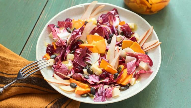 Ein Teller mit Radicchio-Salat, Birnenscheiben, Käsewürfeln, Kürbiskernen, Cranberries und orangefarbener Frucht auf Holztisch.