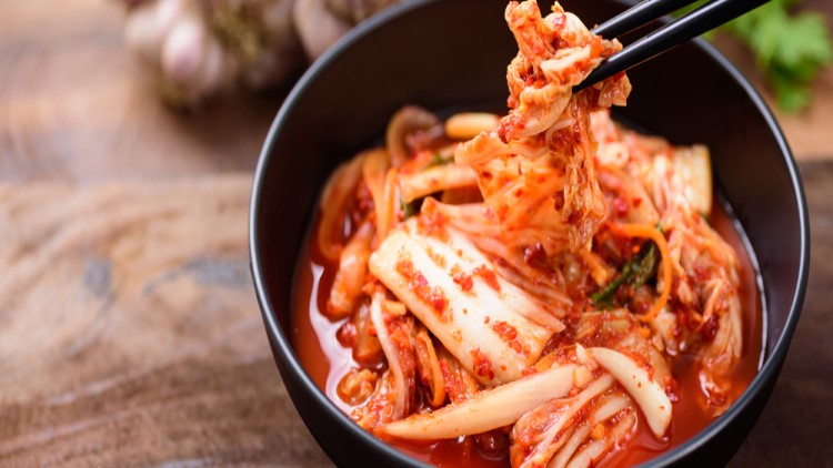 Schwarze Schale mit scharf gewürztem Kimchi, daneben Holzuntergrund und unscharfe Knoblauchknollen im Hintergrund.