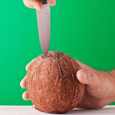 Eine Hand hält eine braune Kokosnuss, während ein Messer von oben in die Schale gedrückt wird, Hintergrund ist grün.