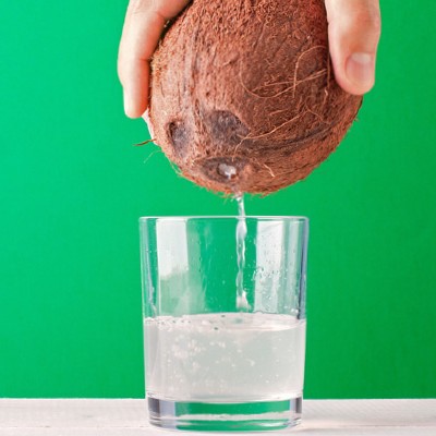 Eine Hand hält eine Kokosnuss, aus der klarer Saft in ein halb gefülltes Glas vor einem grünen Hintergrund fließt.