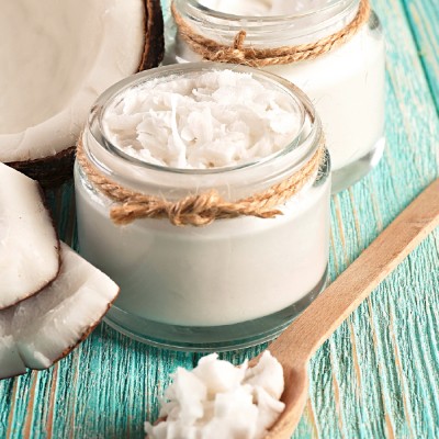 Kleine Gläser mit Kokosöl und Kokosraspeln, dekoriert mit Juteband, neben frischen Kokosnussstücken auf türkisfarbenem Holz.