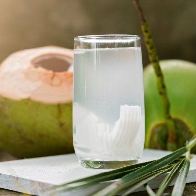 Ein Glas mit klarem Kokoswasser und Fruchtfleischstücken steht vor zwei grünen Kokosnüssen auf einem weißen Untergrund.