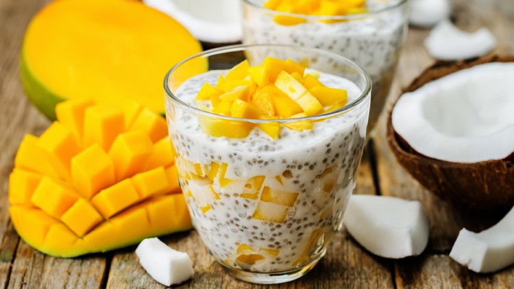 Zwei Gläser mit Chia-Pudding, frischen Mangostücken und Kokosstücken, umgeben von einer halben Mango und geöffneter Kokosnuss.