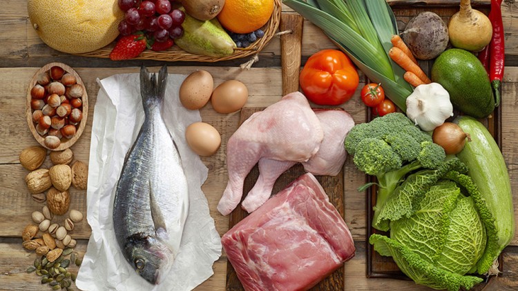 Verschiedene Lebensmittel wie Obst, Gemüse, Nüsse, Fisch, Eier, Hähnchenschenkel und ein Fleischstück liegen auf einem Holztisch.