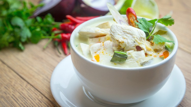 Eine weiße Suppenschale mit Hühnersuppe, Kokosmilch, Gemüse und Koriander auf einem Holztisch, im Hintergrund frische Zutaten.