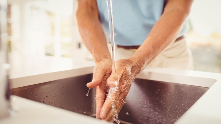 Person wäscht die Hände unter fließendem Wasser in einem modernen Waschbecken in heller Umgebung.