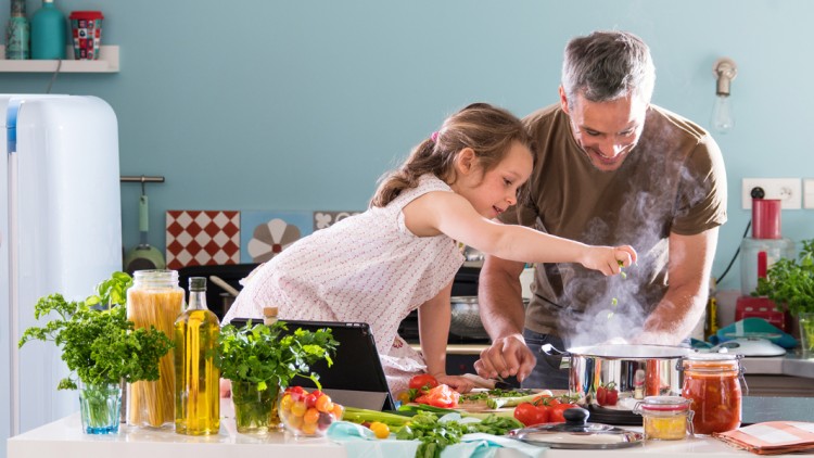 Ein Kind und ein Erwachsener kochen gemeinsam an einem Küchentisch mit frischem Gemüse, Kräutern und einer dampfenden Pfanne.