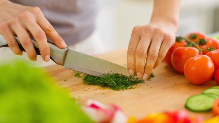 Hände schneiden frische grüne Kräuter mit einem Küchenmesser auf einem Holzbrett, daneben liegen Tomaten und Gurkenscheiben.
