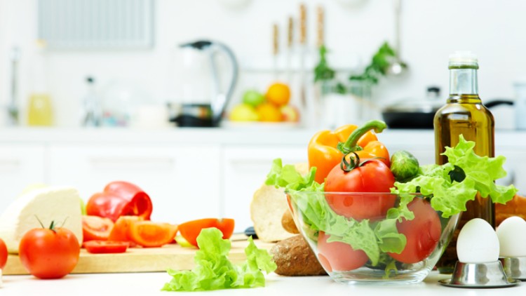 Auf einer Küchenarbeitsplatte stehen eine Glasschüssel mit Gemüse, geschnittene Paprika, Brot, Käse, Eier und eine Ölflasche.