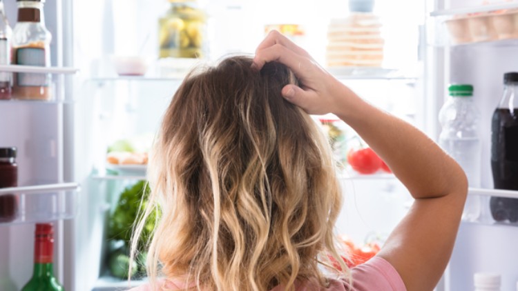 Person von hinten mit schulterlangem Haar blickt in einen offenen Kühlschrank voller Lebensmittel und fasst sich an den Kopf.