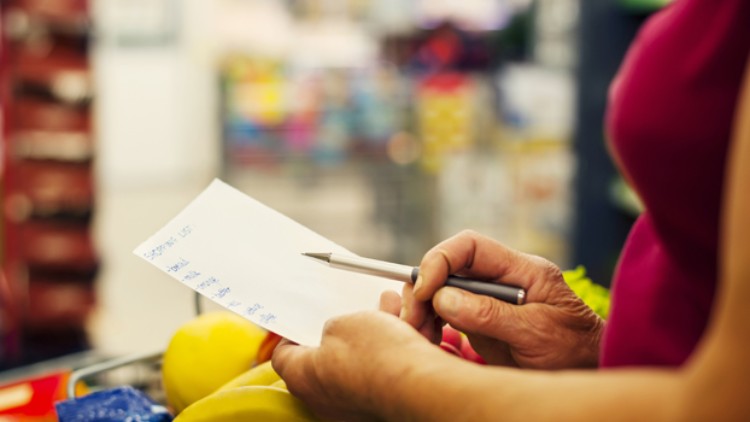 Person hält eine Einkaufsliste und einen Stift, Vordergrund mit gelbem Obst, Hintergrund unscharfer Supermarkt.