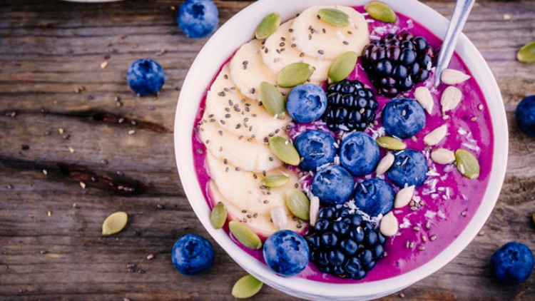 Eine Schale mit pinker Smoothie-Bowl, belegt mit Bananenscheiben, Blaubeeren, Brombeeren, Samen und Kernen auf Holztisch.
