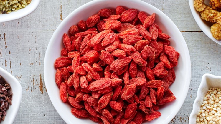 Rote Gojibeeren liegen in einer weißen Schale, umgeben von weiteren Schalen mit Körnern und getrockneten Zutaten.