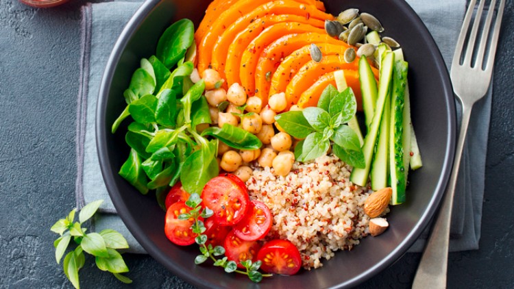 Ein schwarzer Teller mit Quinoa, Kichererbsen, Babyspinat, Tomaten, Gurken, Kürbisscheiben, Kürbiskernen und Kräutern.