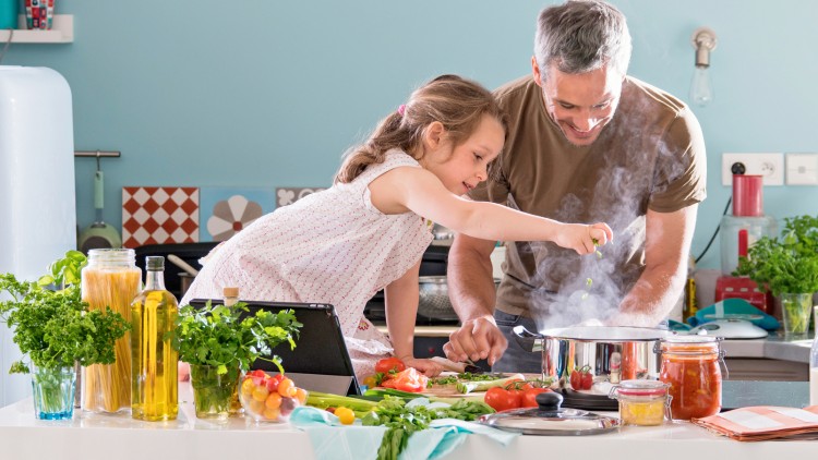 Kind und Erwachsener kochen gemeinsam, mit frischen Zutaten wie Kräutern, Gemüse und Öl auf einer Kücheninsel.