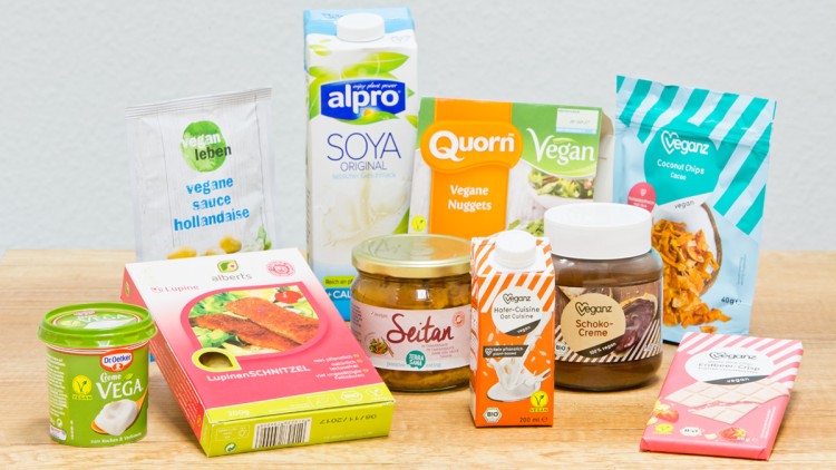 Eine Auswahl an veganen Produkten wie Saucen, Schokolade, Nuggets, Getränken und Aufstrichen steht auf einer Holzfläche.
