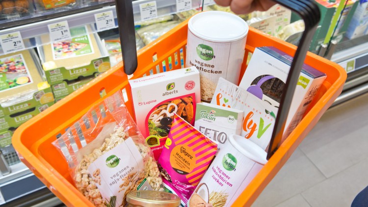 Orange Einkaufskorb mit verschiedenen veganen Produkten, darunter vegane Hefeflocken, Sojafleisch und Seitan, vor einem Kühlregal.