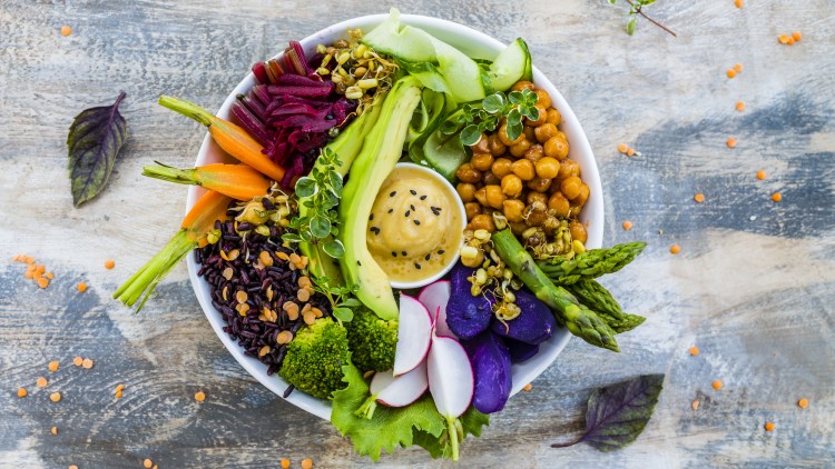 Weiße Schüssel mit Hummus in der Mitte, umgeben von Avocado, Kichererbsen, Gemüse, lila Kartoffeln, Reis und Sprossen.
