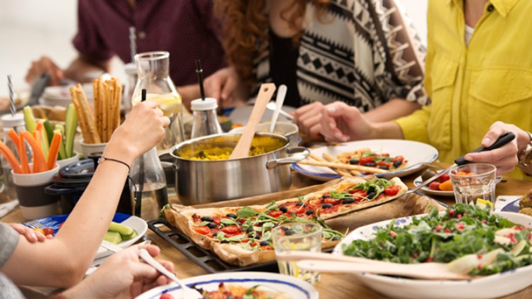 Mehrere Personen sitzen an einem Tisch mit Pizza, Salat, Gemüse, Brotsticks und einem Topf mit gelbem Reis.