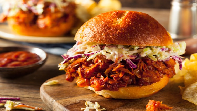 Burger mit geschmortem Gemüse in roter Sauce und Krautsalat auf einem Holzbrett, daneben Chips und eine Schale mit Sauce.