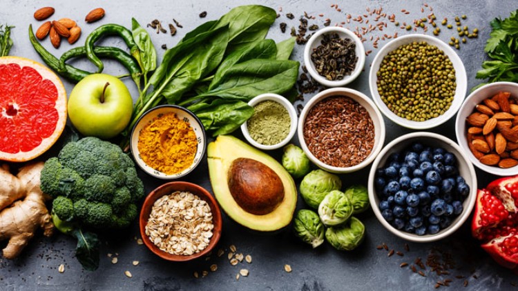 Verschiedene frische Lebensmittel wie Obst, Gemüse, Nüsse, Samen und Gewürze sind auf einem grauen Untergrund arrangiert.