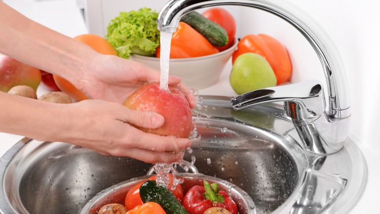 Hände waschen einen roten Apfel unter fließendem Wasser über einem Sieb mit Gemüse wie Paprika, Gurke und Kartoffeln im Spülbecken.