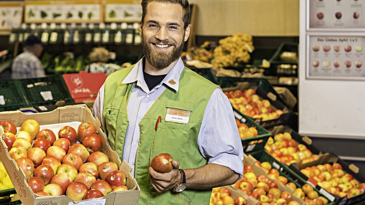 Mitarbeitende Person in grüner Weste präsentiert rote Äpfel in einer Obst- und Gemüseabteilung eines Supermarktes.