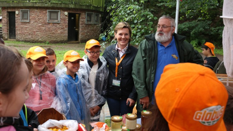 Personen mit orangen GLOBUS-Kappen stehen an einem Tisch mit Honiggläsern und Süßigkeiten in einer grünen, bewaldeten Umgebung.