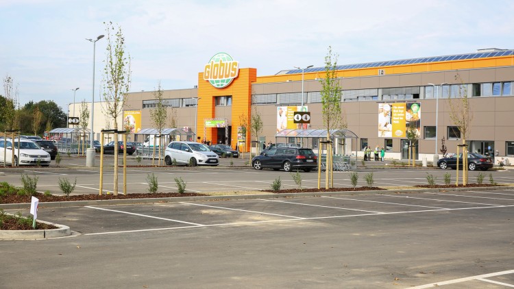 Das GLOBUS Gebäude mit orangefarbenem Eingang, Parkplätzen, Bäumen und Einkaufswagen-Überdachungen im Vordergrund.