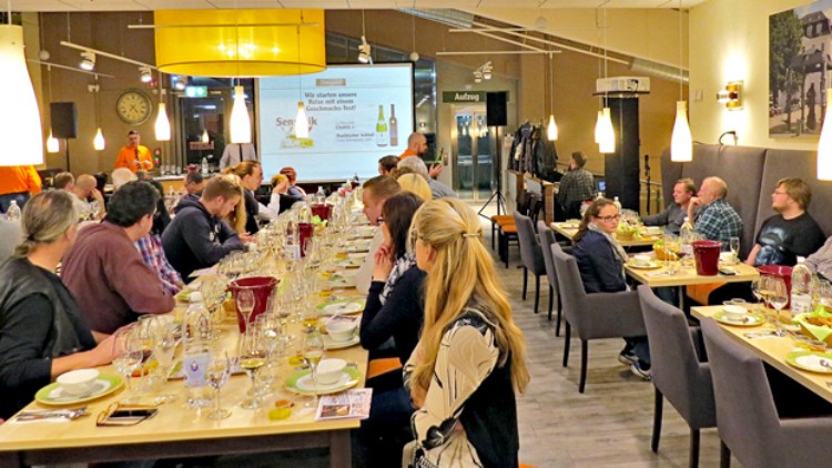 Mehrere Personen sitzen an festlich gedeckten Tischen mit Tellern, Gläsern und Wein im Raum, eine Präsentation ist im Hintergrund.