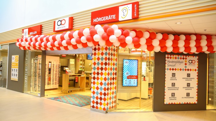 Eingang eines Optikergeschäfts mit roter Ballondekoration, Beschilderung für Brillen und Hörgeräte, sowie Serviceinformationen.