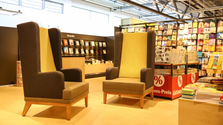 Zwei braun-gelbe Sessel mit Holzbeinen stehen in einer Buchabteilung mit Regalen, Bestseller-Büchern und roten Aktionsschildern.