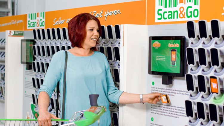 Person entnimmt ein Handscanner-Gerät an einer orange-weißen Scan & Go Station mit zahlreichen Fächern und grüner Anleitung.