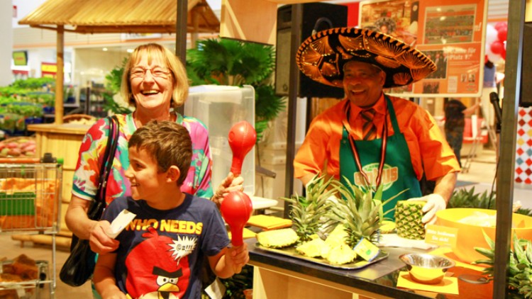 Ein Verkaufsstand mit Ananasstücken, tropischer Dekoration und einem Mitarbeitenden in orangefarbener Kleidung mit großem Sombrero.
