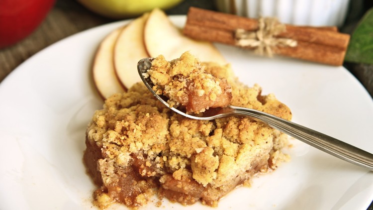 Ein Stück Apfel-Crumble auf einem weißen Teller, im Hintergrund Apfelscheiben und Zimtstangen, ein Löffel nimmt eine Portion.