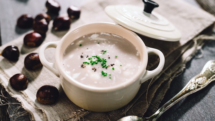 Weiße Keramikschale mit Sahnesuppe, garniert mit gehacktem Schnittlauch und Pfeffer, auf braunem Tuch, Kastanien liegen daneben.