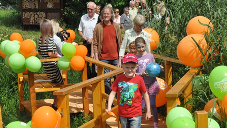 Menschen gehen über einen Steg mit orange-grünen Luftballons, umgeben von hohen Pflanzen und einer Insektennistwand.