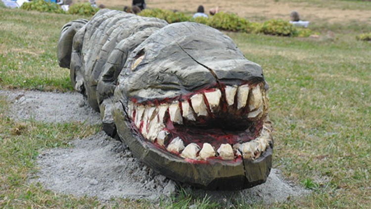 Holzskulptur eines Krokodils mit geöffnetem Maul und sichtbaren Zähnen liegt auf einer Wiese mit unscharfem Hintergrund.