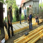 Mehrere Personen stehen und arbeiten an einem Holzrahmen auf einer Baustelle im Freien mit Gebäude und Bäumen im Hintergrund.