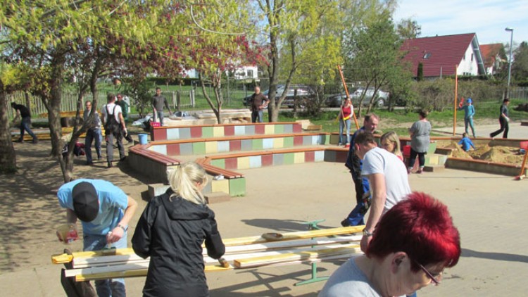 Mehrere Personen arbeiten gemeinsam auf einem Spielplatz, streichen Holzbretter und säubern den Bereich bei sonnigem Wetter.