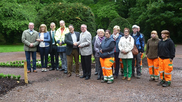 Eine Gruppe von Personen steht in einem grünen Park auf einem Weg, einige tragen orange Arbeitskleidung.