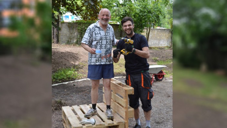 Eine Person steht auf Holzpaletten, eine andere hält eine gelbe Bohrmaschine, im Hintergrund eine Schubkarre und Bäume.