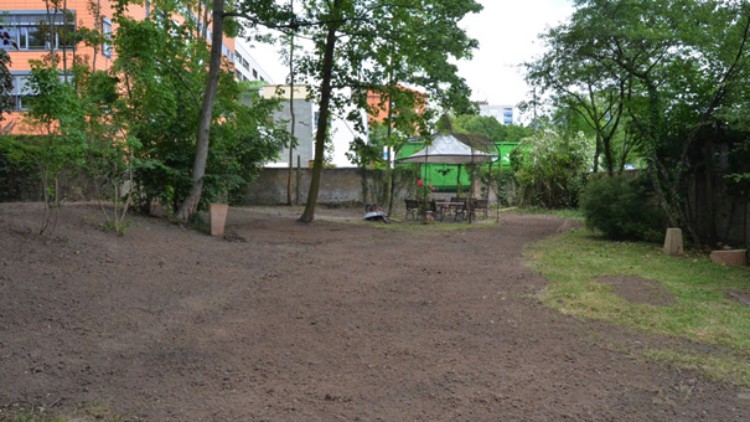Gartenfläche mit frisch aufgelockerter Erde, grünen Bäumen, einem weißen Pavillon und einem Tisch mit Stühlen im Hintergrund.