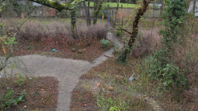 Ein Garten mit unbefestigten Wegen, kahlen Bäumen und überwucherten Beeten in einer herbstlichen Landschaft.