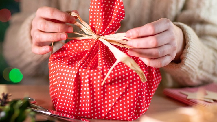 Hände binden eine Schleife aus naturfarbenem Band um ein rotes Geschenk mit weißen Punkten, im Hintergrund bunte Lichter.