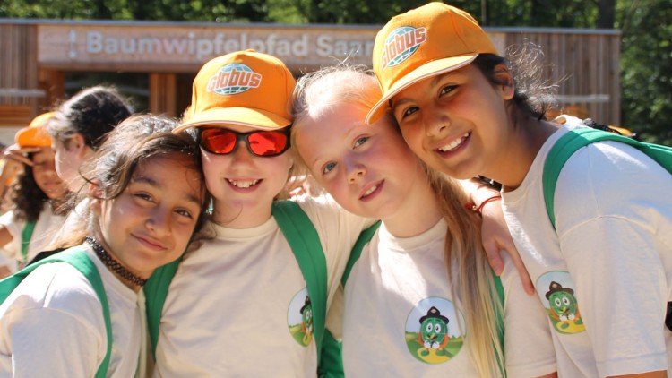 Gruppe von Personen mit orangefarbenen GLOBUS-Kappen und grünen Rucksäcken vor einem Gebäude mit der Aufschrift Baumwipfelpfad Saarschleife.