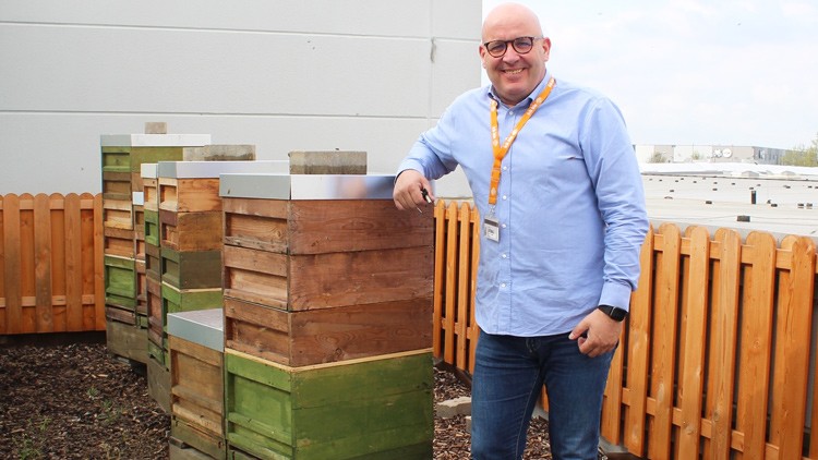 Ein Mann in blauer Kleidung steht neben mehreren Holzbehausungen für Bienen vor einem Holzzaun und einer weißen Wand.