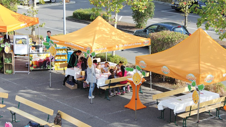 Zwei orange GLOBUS-Pavillons auf einem Parkplatz mit Tischen und Bänken, an denen Kinder und Erwachsene sitzen.